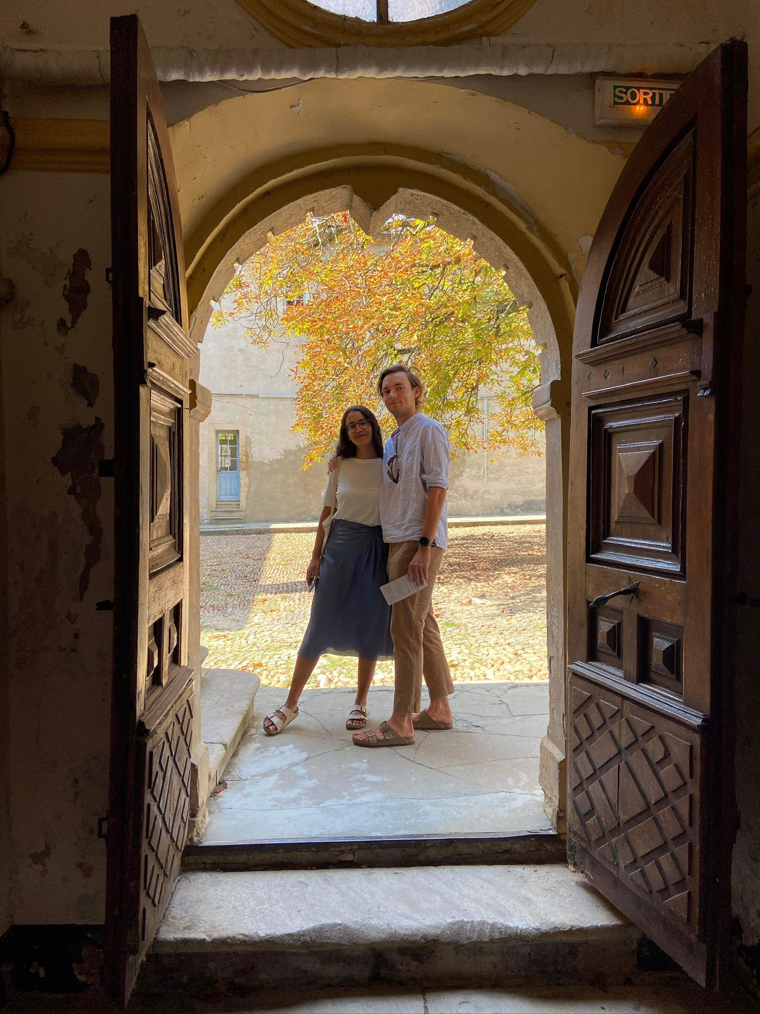 A vintage wooden desk with a mirror in the corner of a room with patterned wallpaper.